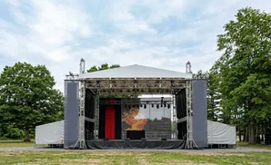 An outdoor stage setup with a white tented roof, trussing, speakers, and a large screen displaying a graphic.