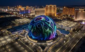 An illuminated spherical building with a swirling, colorful digital display at night, surrounded by city lights and parking lots.