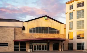 A modern building with stone and light-colored facade, featuring large windows and a prominent green logo on the roof against a sunset sky.