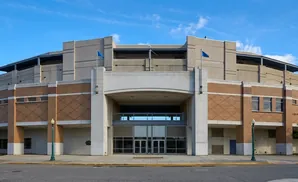 Large brick and concrete building with a wide arched entrance, two blue flags, and glass doors.