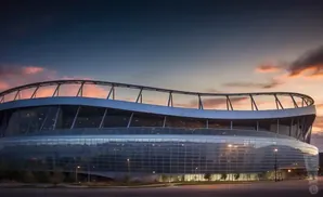 A modern stadium with a glass facade and curved roof against a colorful sunset sky.