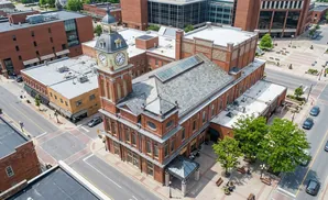 Aerial view of a brick clock tower building in a downtown area with surrounding businesses and street.