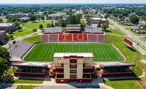 Aerial view of a football stadium with red and gold seating, a green field, and a press box.