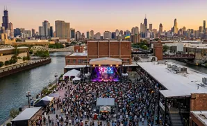 An aerial view of an outdoor concert with a band performing on a stage in front of a large crowd, with a city skyline in the background.