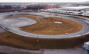Aerial view of a race track with brown grass and a large warehouse in the background.