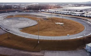 Aerial view of a race track with brown grass and a large industrial building in the background.