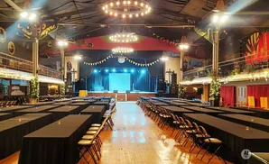 A large hall with rows of black-covered tables and chairs, a stage at the front, and a disco ball hanging from the ceiling.