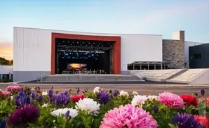 Outdoor amphitheater with a stage, seating, and a foreground of colorful flowers under a bright sky.