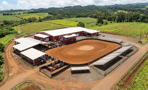 Aerial view of a rodeo arena with red buildings, bleachers, and cattle pens, surrounded by green fields and trees.