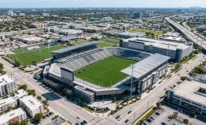 Aerial view of a modern soccer stadium with a green field, surrounded by urban development and a highway.