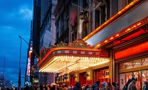 A brightly lit theater marquee shines at dusk with a crowd of people below.
