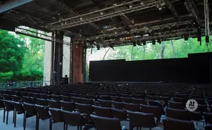 Outdoor amphitheater with rows of dark chairs facing a stage backed by black curtains and surrounded by trees.