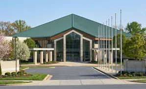 A modern building with a green, pitched roof and large glass entrance, surrounded by trees and flagpoles.