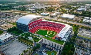 Aerial view of a baseball stadium with red seating and a green field, surrounded by urban development and trees.