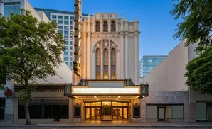 Historic theater with ornate facade and illuminated marquee, flanked by modern buildings and trees.