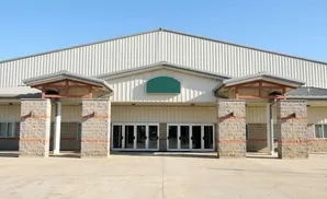 Large, light-colored metal building with a central entrance featuring double glass doors flanked by brick columns.