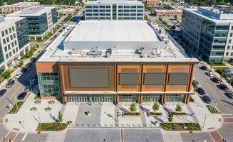 Aerial view of a modern building complex with large screens on its facade, surrounded by trees and parking lots.