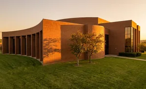 Brick building with curved facade and tall windows, bathed in warm sunset light, with trees and a manicured lawn.