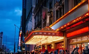 A theater marquee glows with neon lights under a twilight sky, with a crowd gathered below.