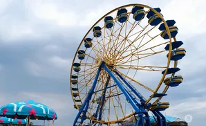 A yellow and blue Ferris wheel stands tall against a cloudy sky, with a patterned umbrella in the foreground.