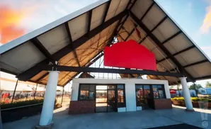 A red, cut-out sign hangs on the underside of a wide, white-tiled, A-frame roof supported by thick white columns.