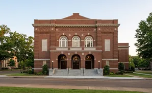 Brick building with arched doorways and windows, with trees and a street in front.
