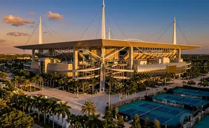 An aerial view of a large stadium with a unique roof design, surrounded by palm trees and tennis courts.