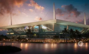 Modern stadium with glass facade and palm trees at sunset, reflected in the water.