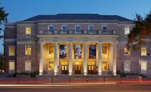 A grand brick building with a portico and large banners is lit at dusk, with car light trails in the foreground.