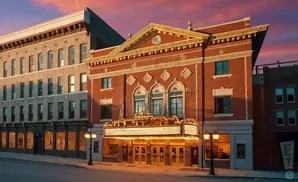 An ornate, red brick theater with a marquee and lights, set against a colorful sunset sky.