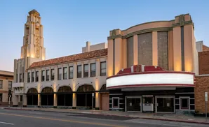 Art deco theater with ornate tower and marquee glows at dusk under a clear blue sky.