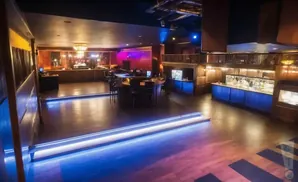 A spacious, dimly lit nightclub with a central bar, raised seating areas, and blue accent lighting on the floor.