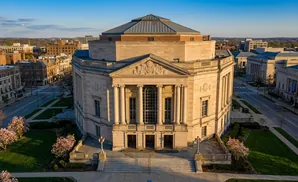 Aerial view of a large, ornate sandstone building with a domed roof, flanked by other classical buildings and city streets.