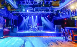 A dimly lit bar stage with a drum set and piano under blue spotlights.