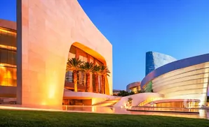 Modern buildings with curved glass and beige stone facades, illuminated at dusk with palm trees and a clear blue sky.