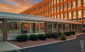 Sunset over a brick building with a glass-enclosed walkway and manicured bushes.