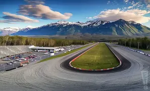 A race track with a grandstand and parking lot, set against a backdrop of snow-capped mountains and trees.