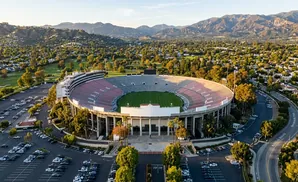 Aerial view of a large, circular stadium with a green field, surrounded by trees and mountains.
