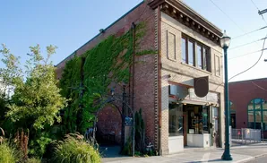 A brick building with ivy, an arched entrance, and a streetlamp stands on a sidewalk under a clear blue sky.