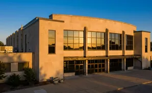 Modern tan stone building with large windows reflecting a sunset under a clear blue sky.
