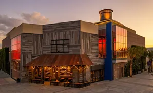A modern building with rustic wooden siding and large windows reflecting a sunset, featuring a covered outdoor dining area with string lights.