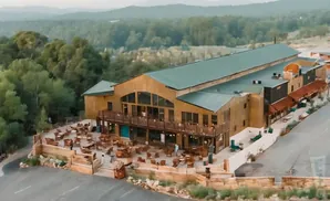 An aerial view of a large, rustic building with a green roof, surrounded by trees and a patio with outdoor seating.