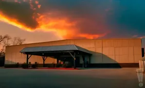 A long, light-colored building with a covered entrance is lit by a dramatic sunset with bright orange and red clouds.