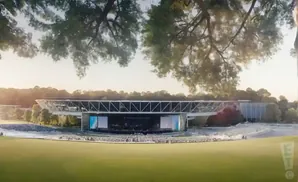 Outdoor amphitheater with a stage, audience seating, and trees in the foreground under a sunset.