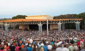 An outdoor amphitheater with a large audience seated on tiered rows facing a stage structure.
