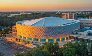 Aerial view of a circular arena with a patterned facade and a large roof, set against a sunset sky.