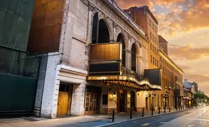 Golden hour light illuminates a grand theater entrance with a marquee and balcony, facing a city street.
