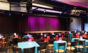 Small tables with bright tablecloths and glowing centerpieces are set up in front of a stage with a purple curtain and microphone.