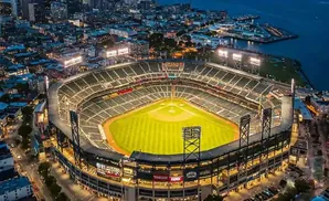 Aerial view of a baseball stadium at dusk, with the city lights and bay in the background.