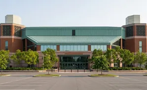 A modern brick building with a green metal roof and large windows, surrounded by trees and a parking lot.